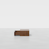 A low, rectangular wooden table with a single drawer partially open, placed on a smooth, light gray floor against a plain white background.