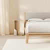 Minimalist bedroom with a light wood bed frame and neutral bedding. A small wooden nightstand holds a carafe, glass, and books. Sheer curtains and a pale rug complete the calm, airy space.