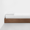 A minimalist bed with a wooden frame, white bedding, and a single white pillow, positioned against a plain white wall and set on a light gray floor.
