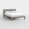 A modern wooden bed frame with a gray upholstered headboard, dressed in white bedding and two white pillows, placed in a minimal, bright room with light-colored flooring and walls.
