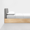 A minimalist bed with a light wooden frame, gray upholstered headboard, and neatly made white bedding, positioned against a white wall on a gray floor.