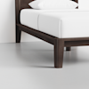 A close-up of a bed with a dark wooden frame, white mattress, and white pillow, shown against a plain light-colored wall and floor.