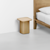 A minimalist wooden nightstand with a smooth, square top sits next to a matching wood bed frame and white bedding in a bright, modern room with light gray flooring.