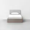 A neatly made single bed with a light gray upholstered headboard, white bedding, and a light wood frame, placed against a plain white wall in a minimal, empty room with a gray floor.