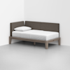 A minimalist daybed with a light wood frame, white mattress, single white pillow, and brown cushioned backrest set against two walls in a corner of a bright, empty room.