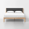 A modern bed with a light wood frame, charcoal gray upholstered headboard, white bedding, and two white pillows, set against a plain white wall and gray floor.