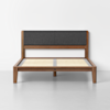 A modern wooden bed frame with a gray upholstered headboard and wooden slats, set against a plain white wall and gray floor.
