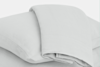 A neatly folded light grey bedsheet is draped over two stacked pillows, all made of soft fabric, against a plain white background.
