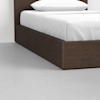 A close-up view of the corner of a wooden bed frame with a white mattress, neatly made with white sheets and a pillow, on a smooth light gray floor.