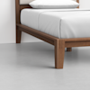 A close-up view of a wooden bed frame with clean lines and tapered legs, featuring white bedding and a white pillow, against a light gray floor and plain white wall.