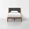 A single bed with a wooden frame and legs, a dark upholstered headboard, white bedding, and a single white pillow, set against a plain white wall and floor.