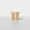 A small, minimalist wooden stool with a light natural finish, featuring a square seat and four wide, flat legs, placed on a smooth, light gray floor against a plain white background.