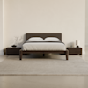 Minimalist bedroom with a dark wooden bed frame, gray upholstered headboard, white bedding, matching wooden nightstands, a small plant, a lamp, and a neutral rug on a light-colored floor.