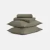 Two olive green pillows stacked on top of each other with folded bedsheets and pillowcases placed neatly on top, all set against a plain white background.
