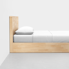 A minimalist wooden bed with a light finish is shown against a white wall. The bed is neatly made with white bedding and a single pillow, placed on a gray concrete floor.