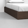A close-up view of a modern bed with a dark wooden platform base, white bedding, and a light gray headboard, set on a light gray floor.