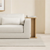 A minimalist living room with a light beige sofa, matching pillow, and a wooden side table holding stacked books and a cup, set on a light carpet against a neutral wall.