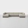 A large, modern, L-shaped sectional sofa with beige upholstery and a low, dark wooden base, set against a plain white background and floor.