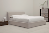 A minimalist bedroom with a beige upholstered bed, white bedding, a dark wooden nightstand with a round lamp, and a shelving unit holding a vase and decorative items against a light-colored wall and carpet.