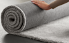 A hand rolls up a thick, soft, gray carpet on a flat surface.
