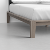 Close-up of the corner of a light wood bed frame with white bedding, showing clean lines and modern design on a smooth, light gray floor.