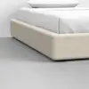 A close-up view of a modern bed with a light beige fabric frame, white bedding, and pillow, set on a smooth, gray floor.