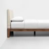 A minimalist bed with a wooden frame, white bedding, a single white pillow, and a light upholstered headboard, set against a plain white wall and floor.
