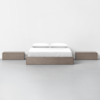 A minimalist bedroom features a wooden platform bed with white bedding and pillows, flanked by two matching wooden nightstands, all set against a plain white wall and gray floor.