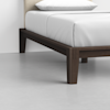 A close-up view of a modern bed with a dark wooden frame and white bedding, showing the corner of the bed against a light-colored wall and floor.