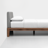 A minimalist bed with a light gray upholstered headboard, white bedding, and a wooden frame, positioned against a white wall on a white floor.