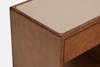A close-up of a wooden piece of furniture with a beige, textured top surface and a smooth brown finish, featuring a drawer and an open lower shelf.
