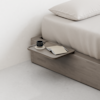 A minimalist bedside table attached to a bed frame holds a black coffee cup, a saucer, and an open notebook with a pen. The bed has beige bedding and is set against a plain white wall.