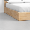 Close-up of the corner of a wooden bed frame with a light finish, white sheets, and pillows on a gray floor.