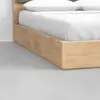 Close-up of the corner of a wooden bed frame with a light finish, white sheets, and pillows on a gray floor.