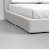 Close-up view of the bottom corner of a modern bed with a light gray upholstered frame and white bedding, set on a smooth, light gray floor.