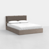 A modern platform bed with a light wood frame and a padded, taupe upholstered headboard, made up with white sheets, pillows, and a duvet; set against a plain white wall and floor.
