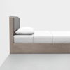 A minimalist wooden bed with a gray padded headboard, white pillow, and neatly made white bedding, positioned against a white wall on a gray floor.