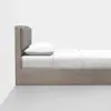 A minimalist wooden bed with a gray padded headboard, white pillow, and neatly made white bedding, positioned against a white wall on a gray floor.