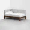 A minimalist daybed with a wooden frame, light gray upholstered backrest, and a white mattress with a single pillow, positioned in the corner of a plain white room.