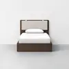 A modern twin bed with a dark wood frame, light beige upholstered headboard, and white bedding, centered against a plain white wall on a light gray floor.