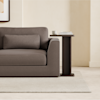 A modern brown sofa with a matching pillow is next to a dark wood side table holding books and a cup, set on a light beige rug in a minimal, neutral-toned room.