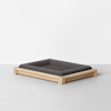 A modern pet bed with a light wooden frame and a dark gray cushioned mattress, set on a light gray floor against a plain white background.