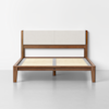 A wooden bed frame with a light beige upholstered headboard, slatted base, and minimalist design, set against a plain white wall on a gray floor.