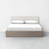 A modern bed with a light wood frame, white bedding, two white pillows, and a beige upholstered headboard, set against a plain white wall and gray floor.