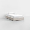 A single bed with a beige upholstered frame, white sheets, and a pillow, set against a plain white wall in a minimalistic, empty room.