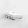 A single bed with a beige upholstered frame, white sheets, and a pillow, set against a plain white wall in a minimalistic, empty room.
