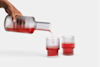 A hand pours red liquid from a ribbed glass carafe into one of two matching ribbed glasses, both partially filled, against a plain white background.