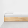 A minimalist wooden bed frame with a white mattress, white sheets, and a white pillow, positioned against a white wall on a light gray floor.