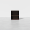 A small, dark brown wooden bookshelf with three shelves, positioned on a plain, light gray floor against a white background.