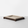 A modern, minimalist pet bed with a light beige cushioned surface and a dark wooden frame, placed on a light gray floor against a plain white background.